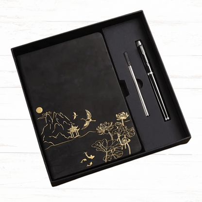 Black notebook with gold designs and a pen in a black box on a white wooden surface