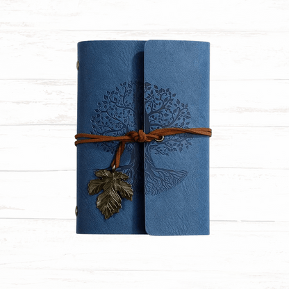 Blue leather-bound notebook with tree design and leaf charm on a white wooden background