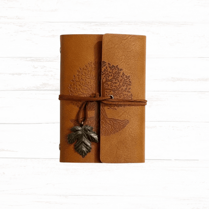 Brown leather journal with tree design and leaf charm on a white wooden surface